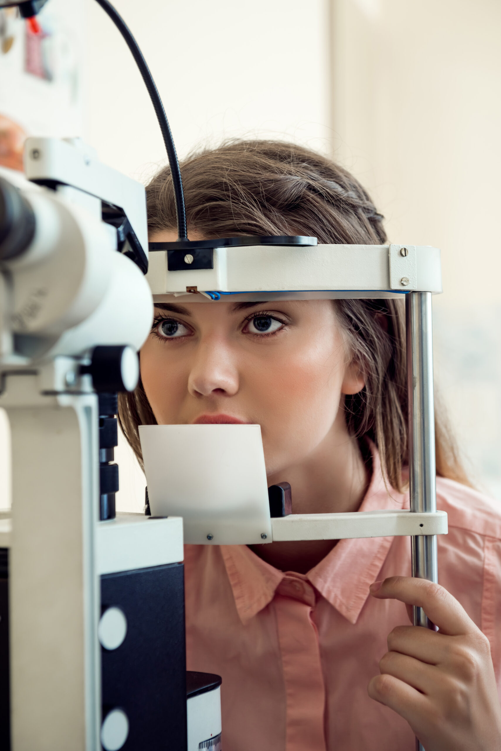 Horizontal portrait of focused european woman testing sight while looking through microbioscope, sitting in specialist office, wanting to pick appropriate glasses to see better