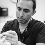 Black and white image of Mr. Daniel Ezra in scrubs, carefully performing a procedure on a patient with a focused expression.
