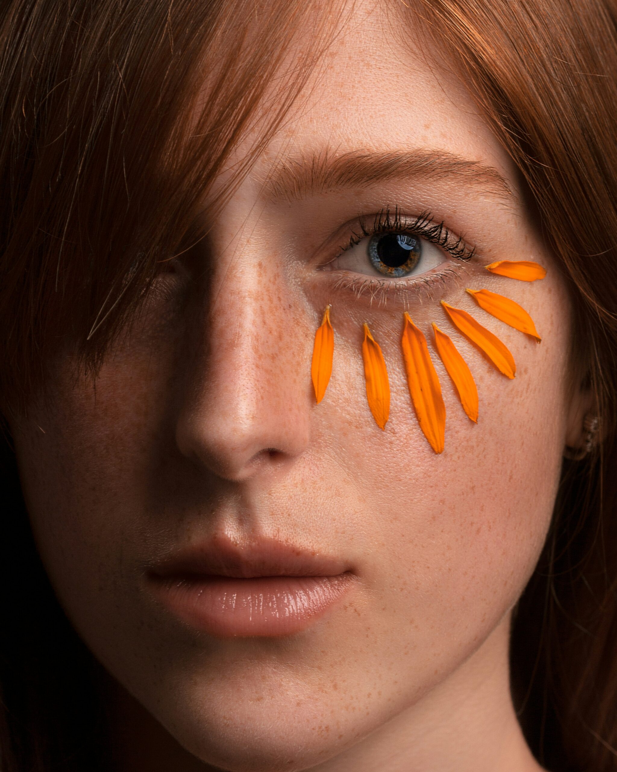 Close-up portrait of a young woman with freckled skin and orange flower petals placed under her eye in a fan-like pattern.