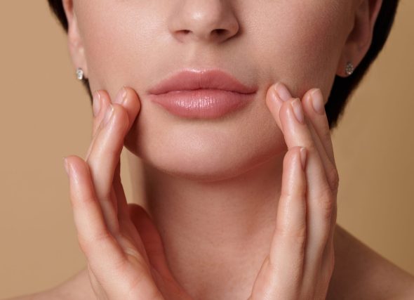 Close-up of a woman gently touching her face near her lips, showing smooth, full lips after lip filler treatment on a beige background