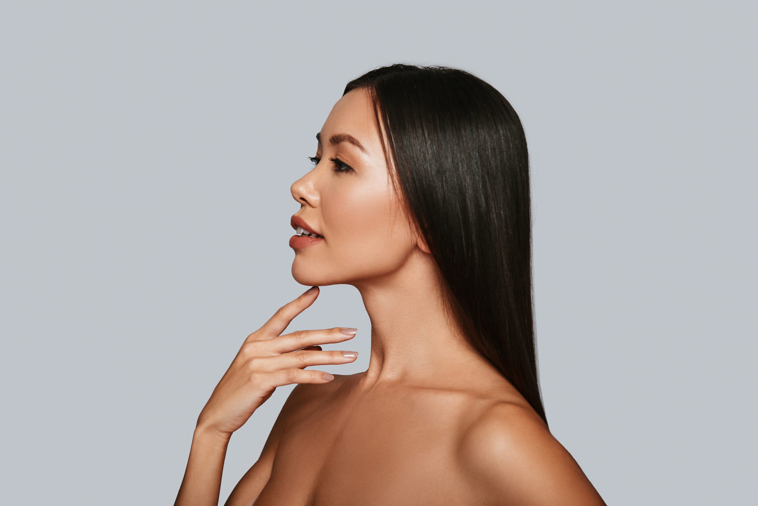 Beautiful young Asian woman smiling while standing against grey background touching her chin