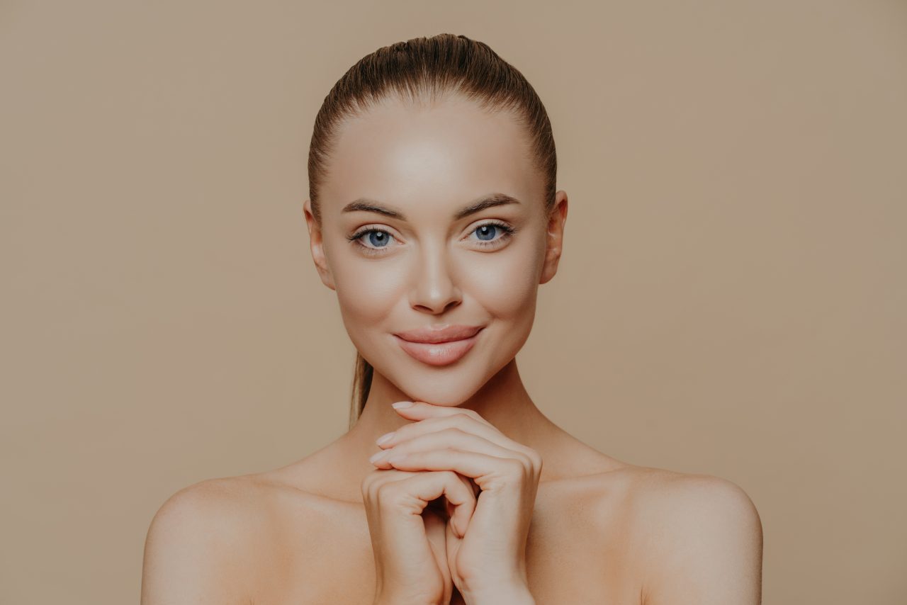 Young pretty woman with beauty skin and combed hair, keeps hands under chin, has healthy complexion after face purification, soft well cared body, isolated over studio wall.