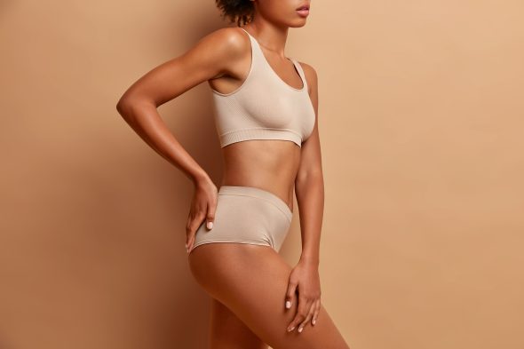 Body of mixed race woman standing in profile keeps hand on buttocks dressed in underwear has perfect shining skin slim figure