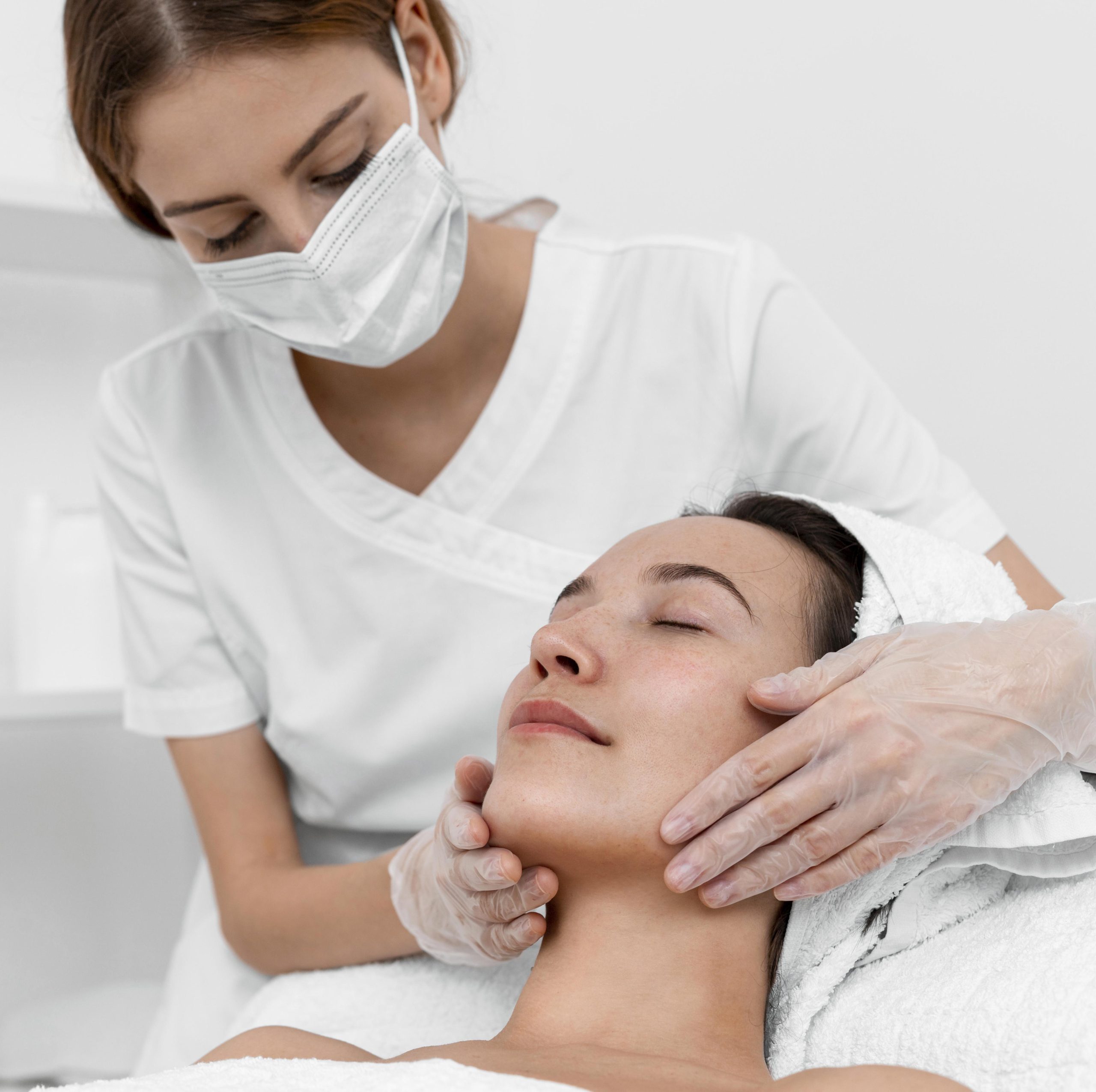 Beautician wearing a face mask and gloves performing a facial massage on a relaxed female client lying with eyes closed and hair wrapped in a towel at a skincare clinic