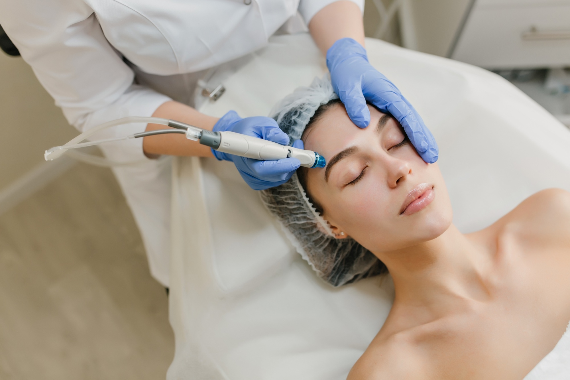 A woman receiving a facial treatment, where a practitioner uses a device on her face, with the patient relaxing and a head covering in place.