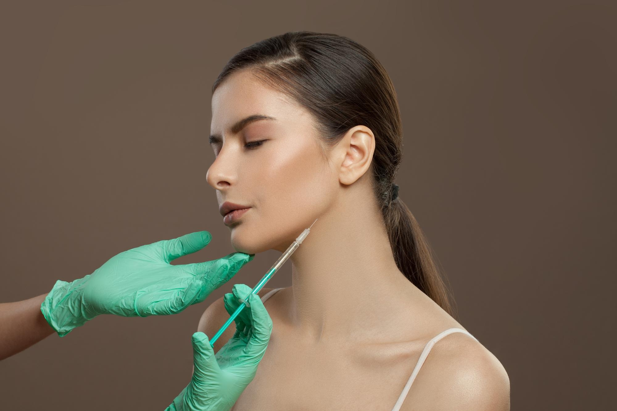 Aesthetician wearing green gloves performing a cosmetic injection along a woman’s jawline for contouring treatment.