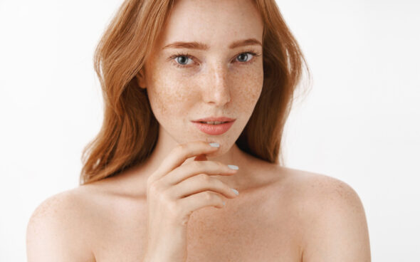 Front-facing portrait of a fair-skinned woman with red hair and freckles, bare shoulders, and a soft expression against a white background.