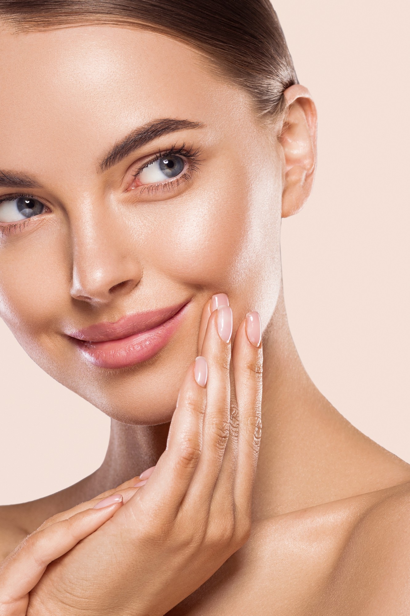 Close-up portrait of a smiling woman with glowing skin gently touching her cheek, showcasing a natural makeup look against a soft neutral background.