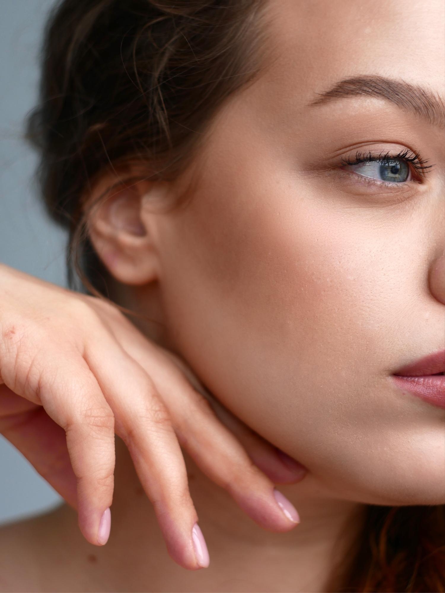 Close-up portrait of a woman with natural makeup resting her hand gently against her face, highlighting smooth skin and soft facial features.
