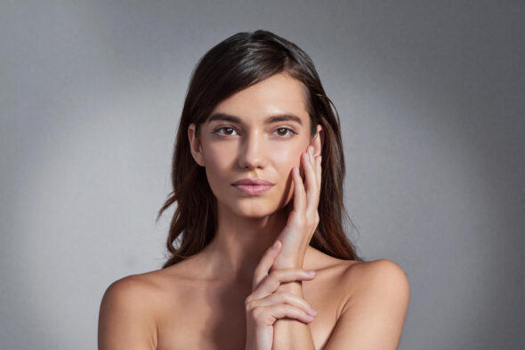 Woman with natural makeup posing against a neutral background, gently touching her face, showcasing smooth and healthy skin.