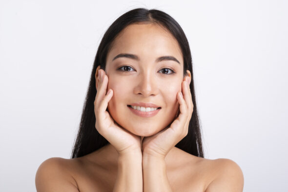 Smiling Asian woman facing the camera with her hands gently framing her face, showcasing clear skin and a natural beauty look against a white background.