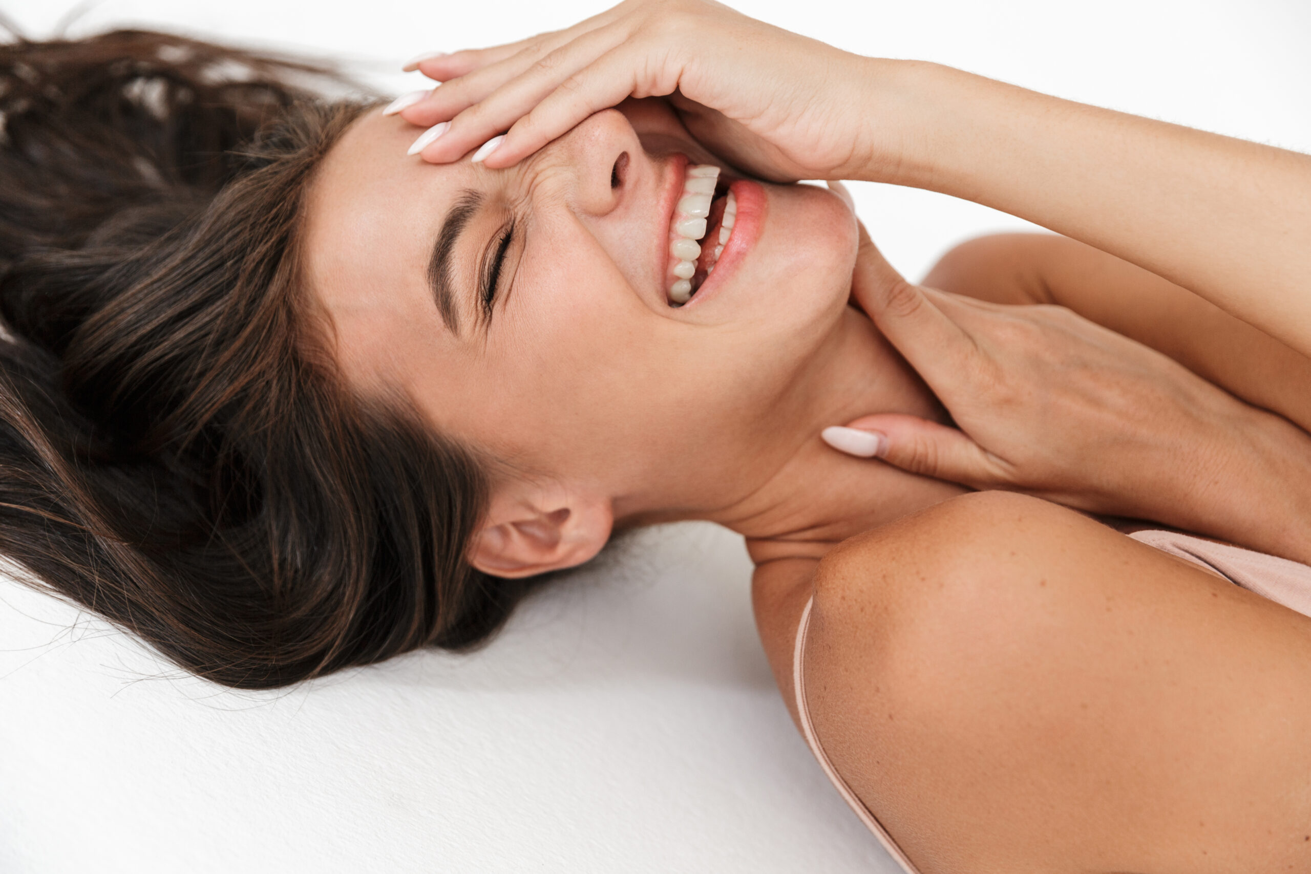 A young woman with smooth, glowing skin laughing with her hand over her face while lying down.