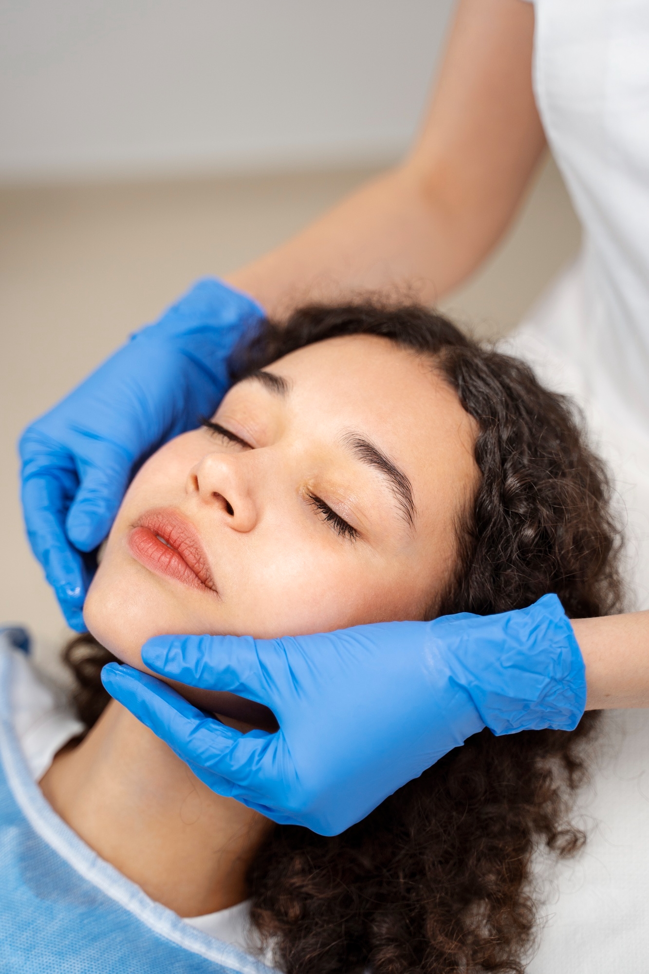 Woman lying down with eyes closed while a practitioner wearing blue gloves gently supports her face during a facial or skincare treatment.