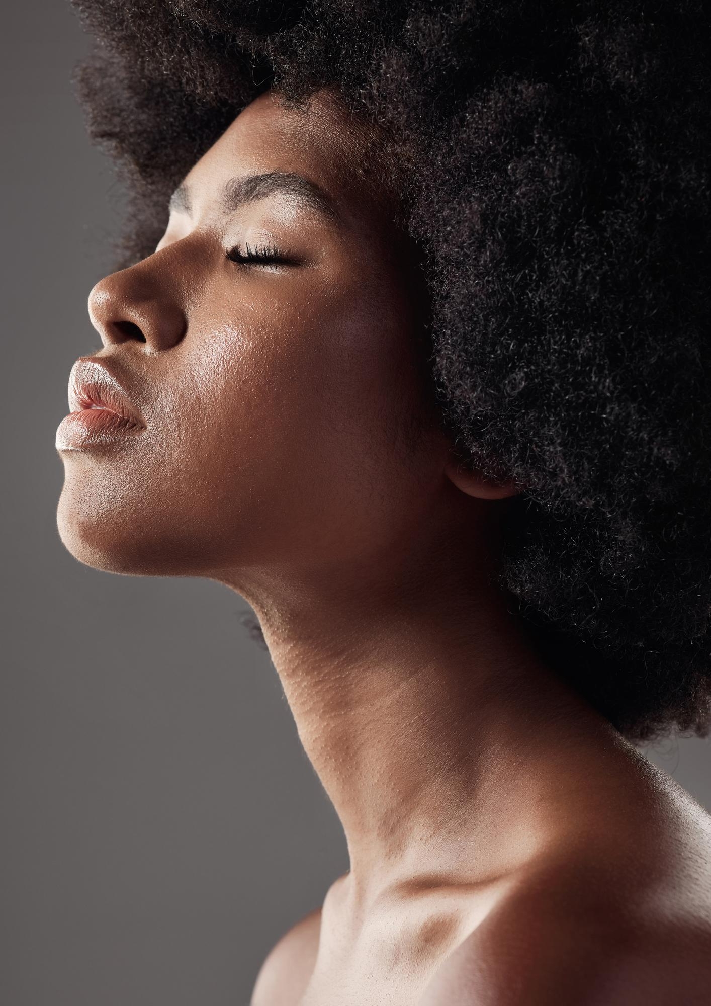 Side-profile portrait of a woman with natural afro hair, eyes closed, head tilted upward, and soft studio lighting highlighting her skin.