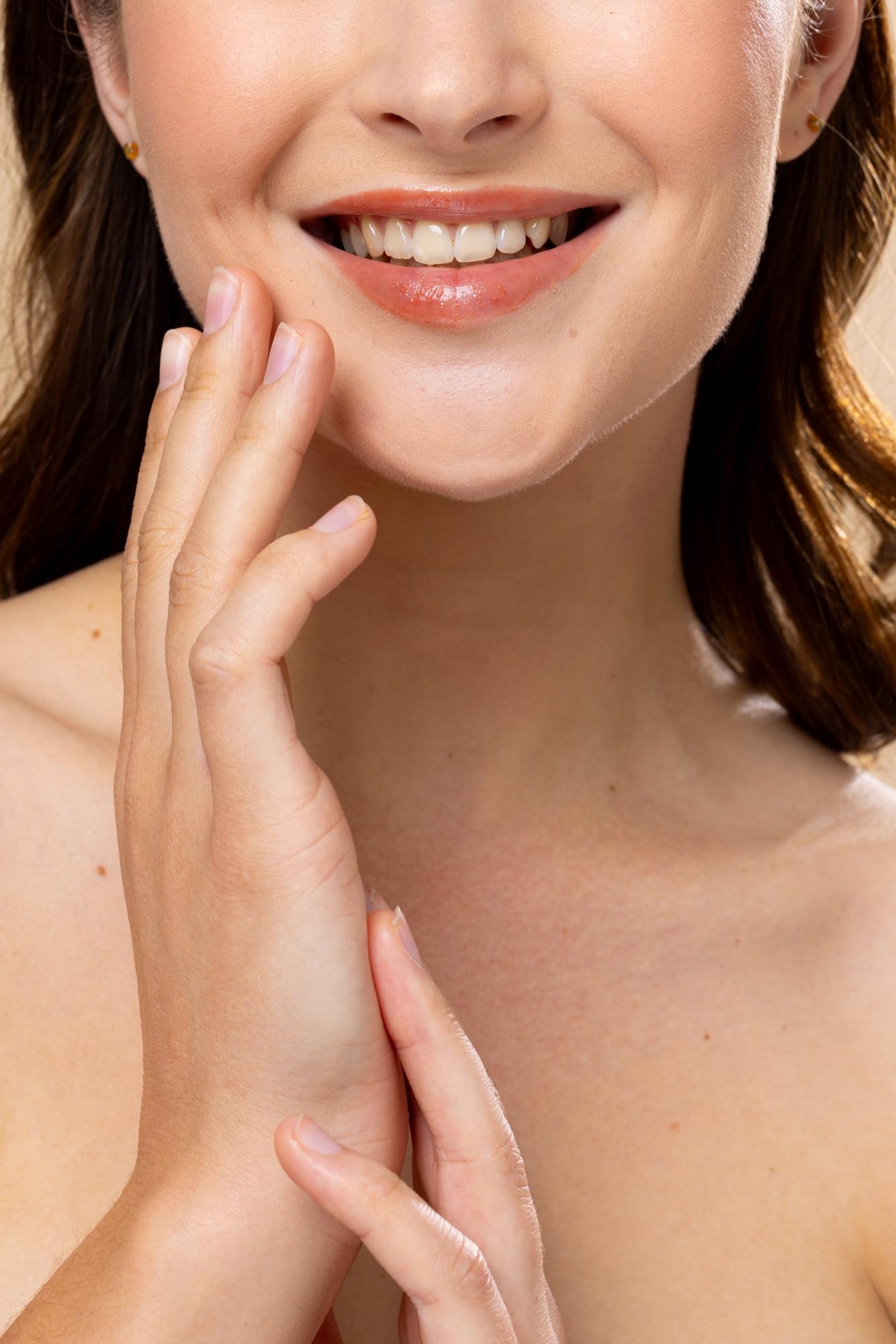 Close-up of a smiling woman with natural lip color, gently touching her face with her hands, highlighting healthy skin and a fresh beauty look.