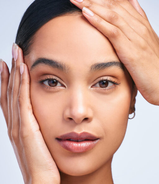 Close-up beauty portrait of a woman looking at the camera with one hand on her cheek and the other resting on her forehead.