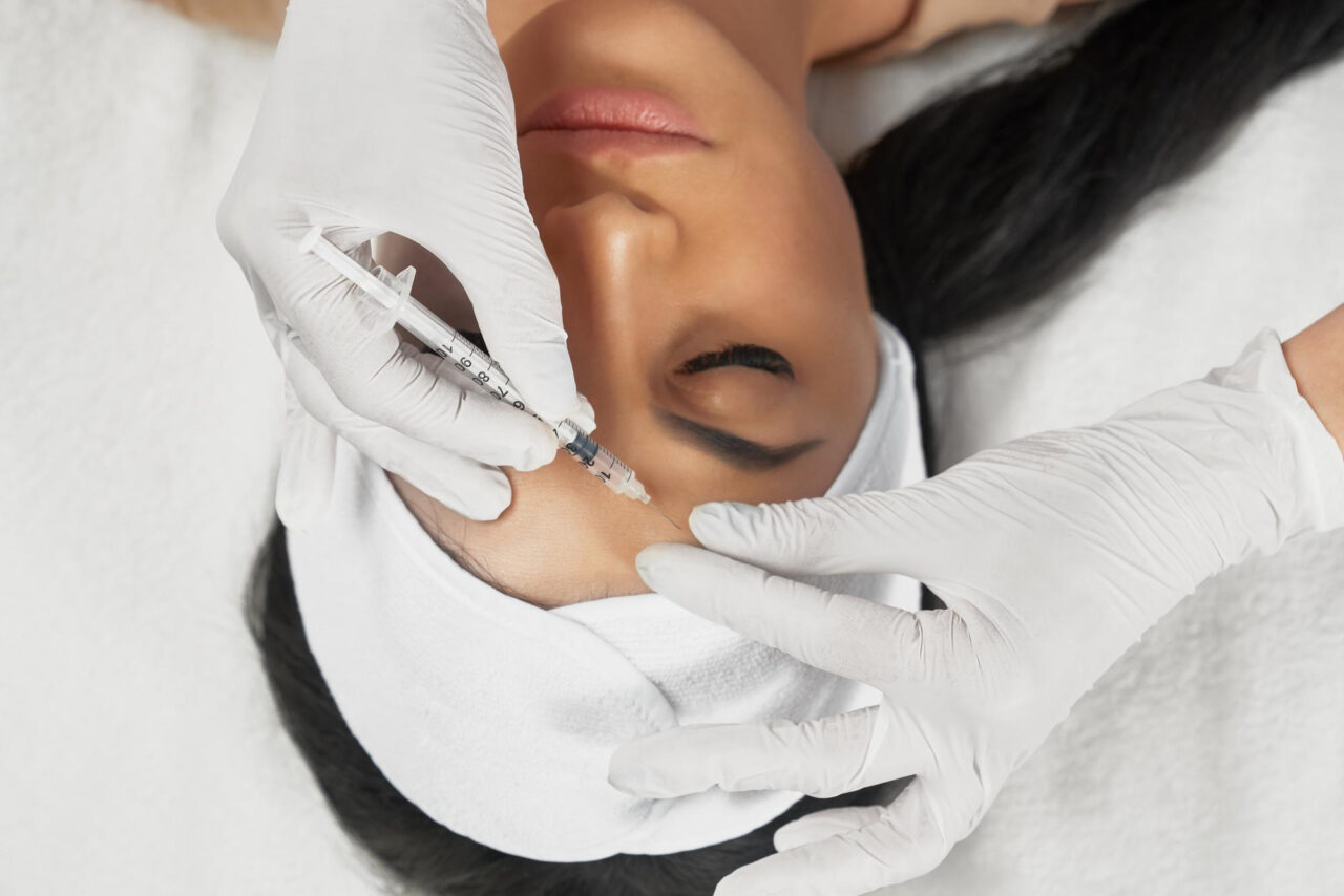 Woman wearing a white headband lies down while white-gloved hands inject a syringe into her cheek area.