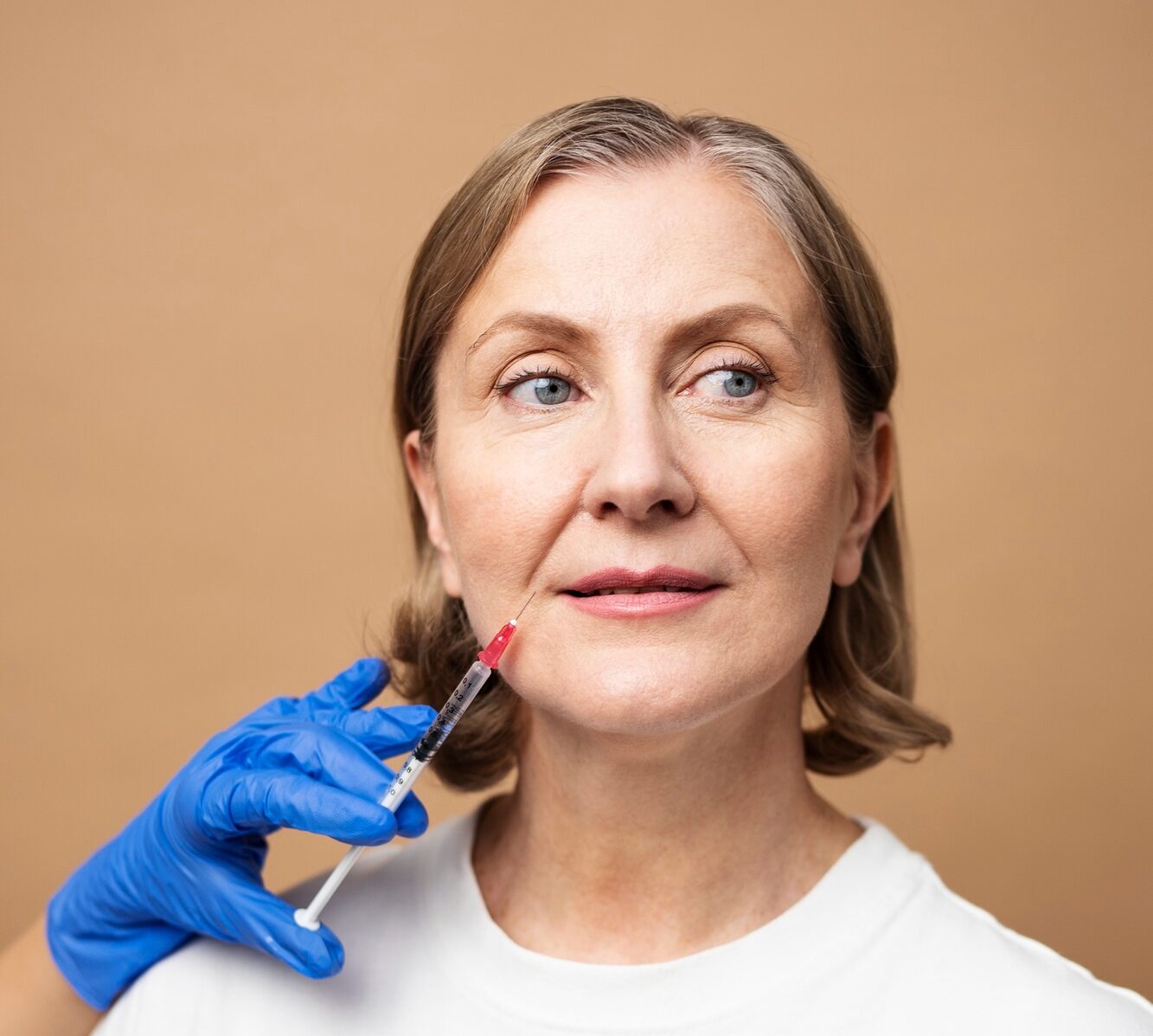 Middle-aged woman looks to the side as a blue-gloved hand holds a syringe near her cheek for a cosmetic injection.