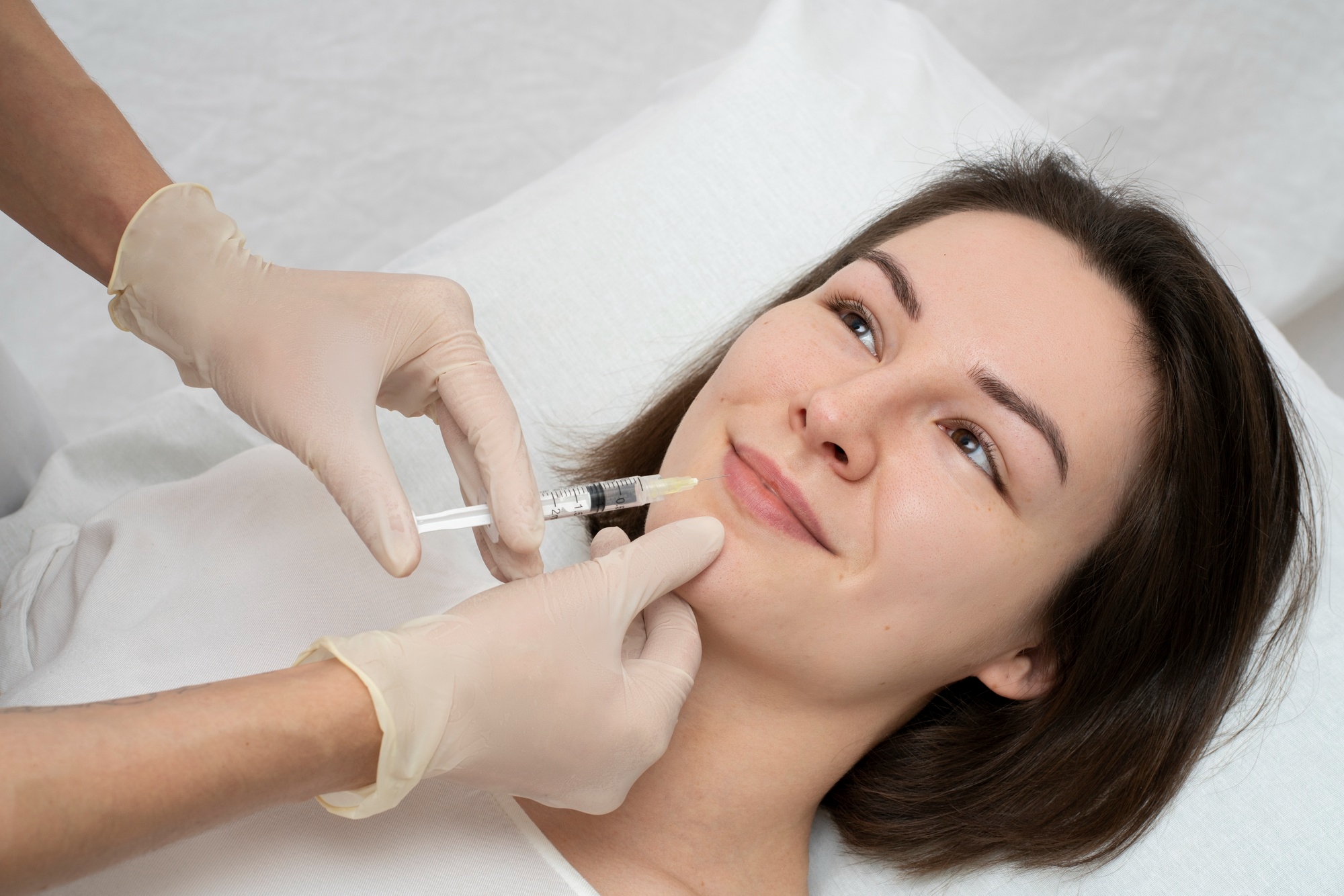 Patient lies on a clinic bed smiling as gloved hands position a syringe at her chin for a cosmetic filler injection.