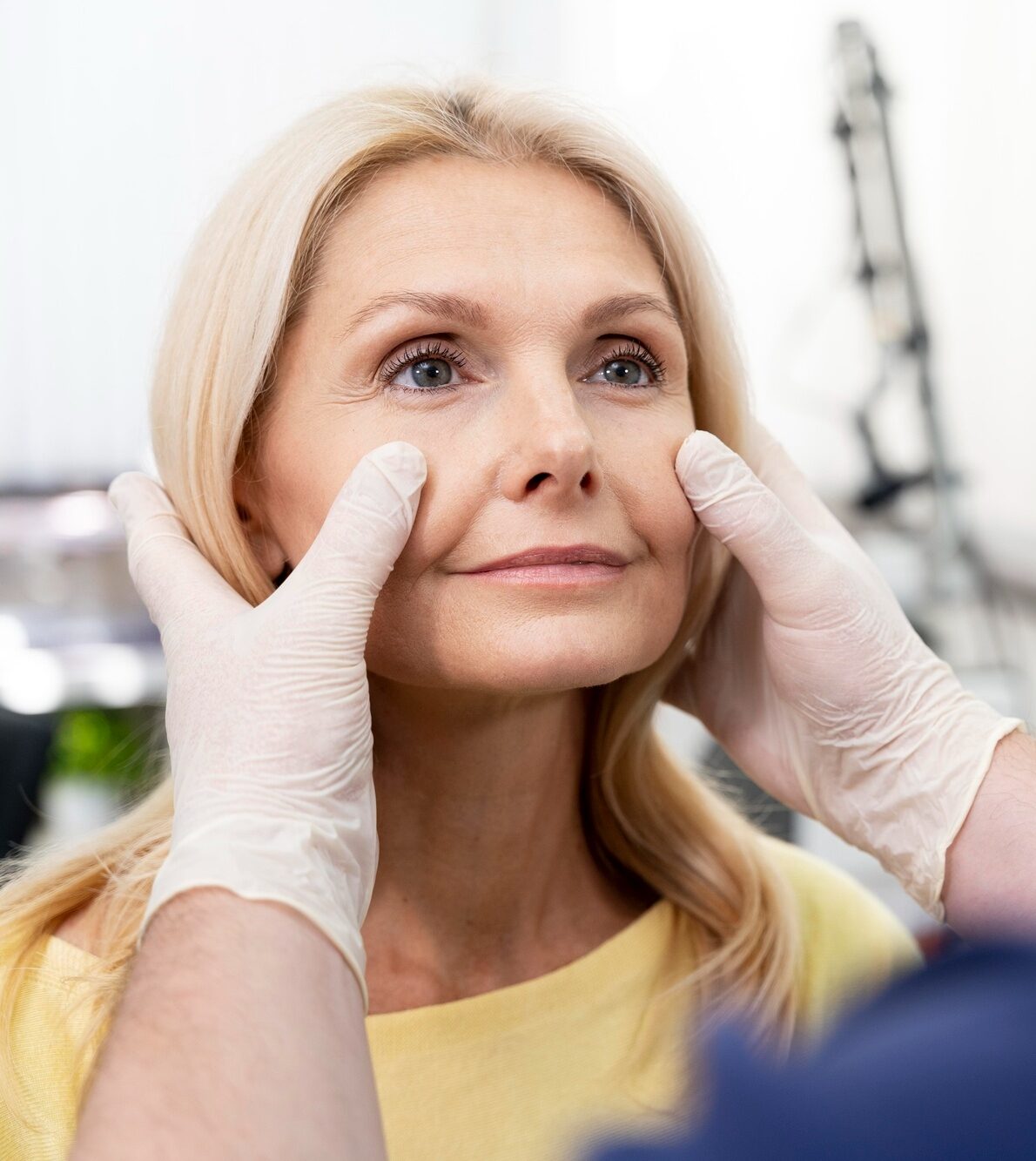 A clinician in white gloves gently presses a blonde woman’s cheeks during a facial assessment in a clinic.