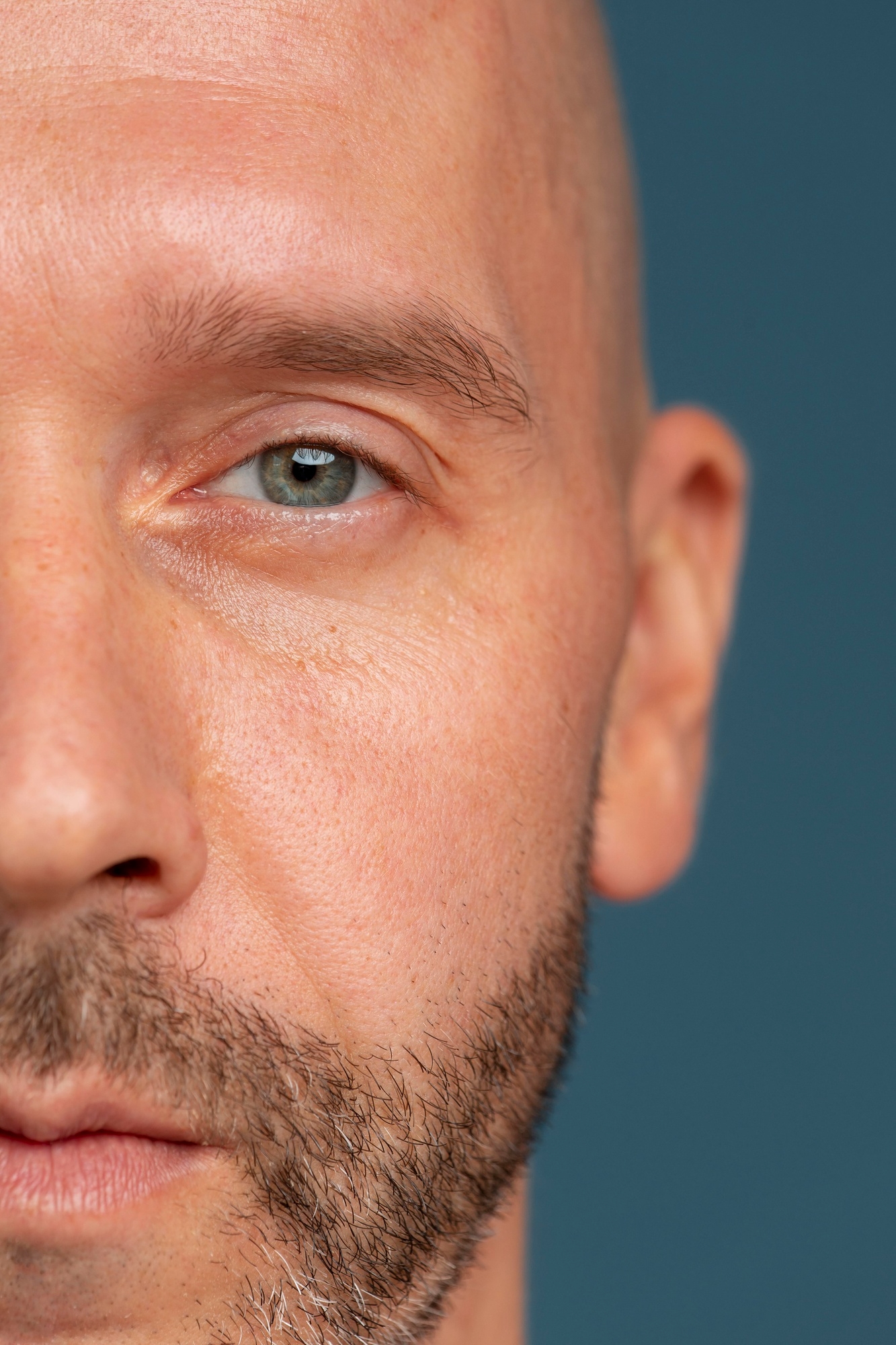 Close-up of the left side of a man’s face showing his eye, eyebrow, and skin texture against a dark teal background.