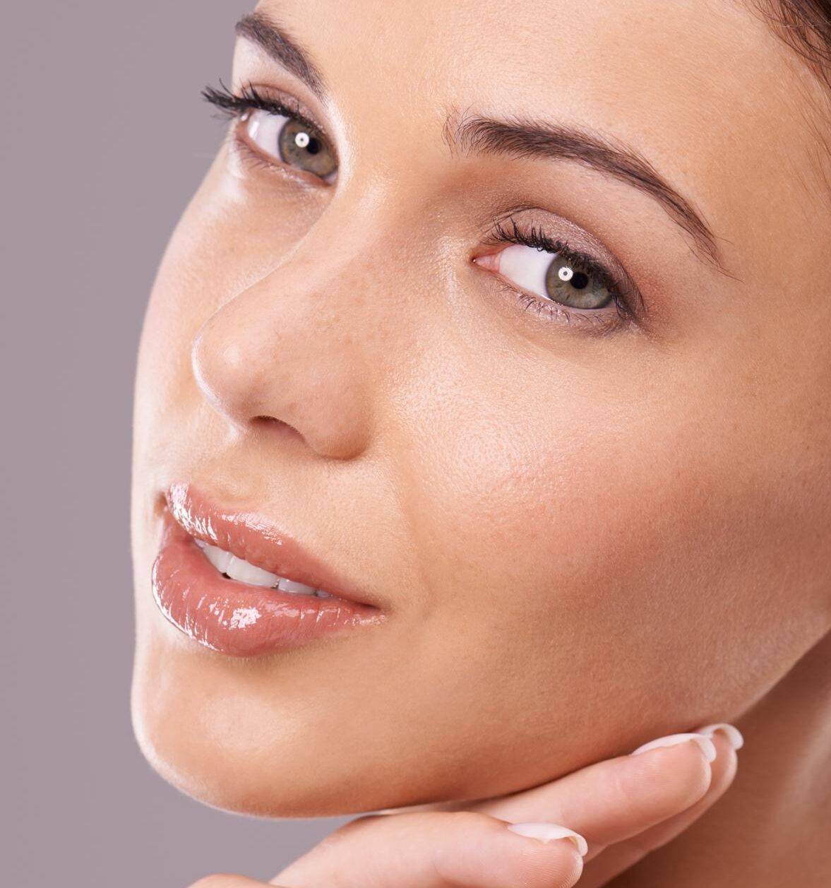 Close-up portrait of a woman with glossy lips resting her manicured hand under her chin against a plain background.