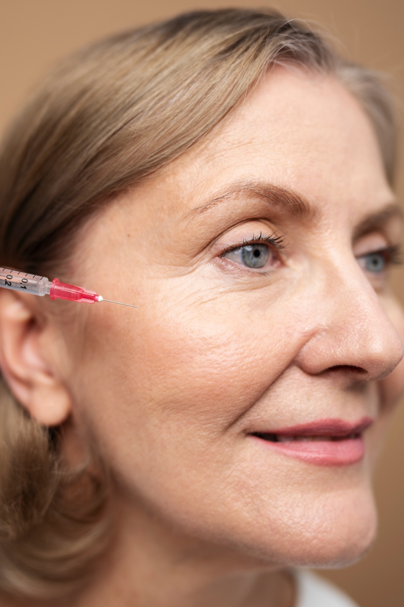 Side-profile close-up of an older woman as a syringe approaches the outer eye area for a cosmetic injection.