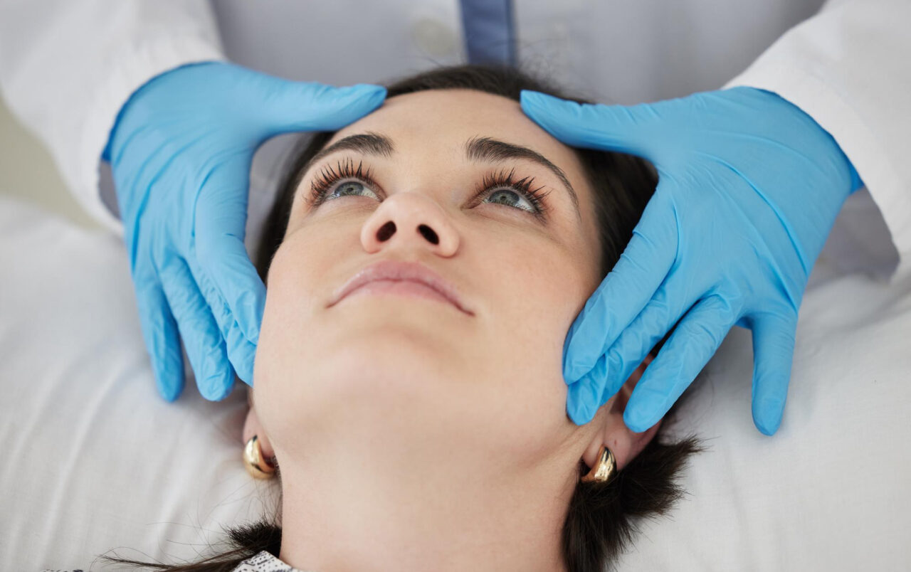 Woman lies on a treatment bed while a practitioner wearing blue gloves examines her face.