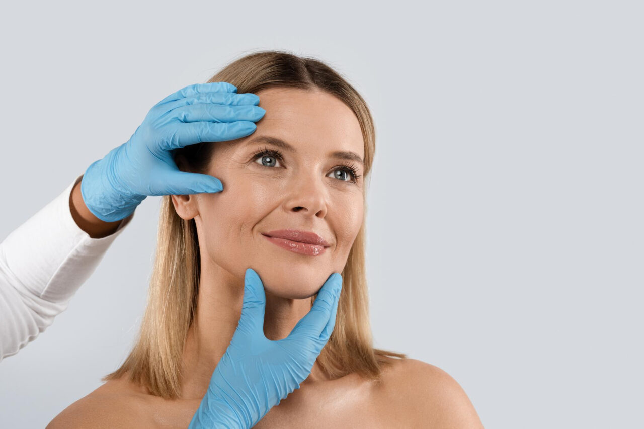 Woman smiling while a practitioner wearing blue gloves examines her face during a cosmetic consultation.