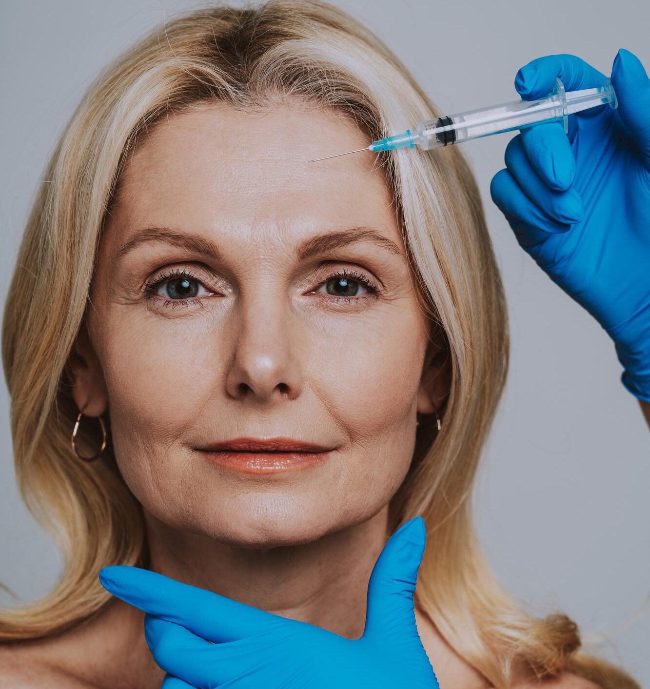 Blue-gloved practitioner steadies an older blonde woman’s chin while bringing a syringe toward her forehead for an injection.