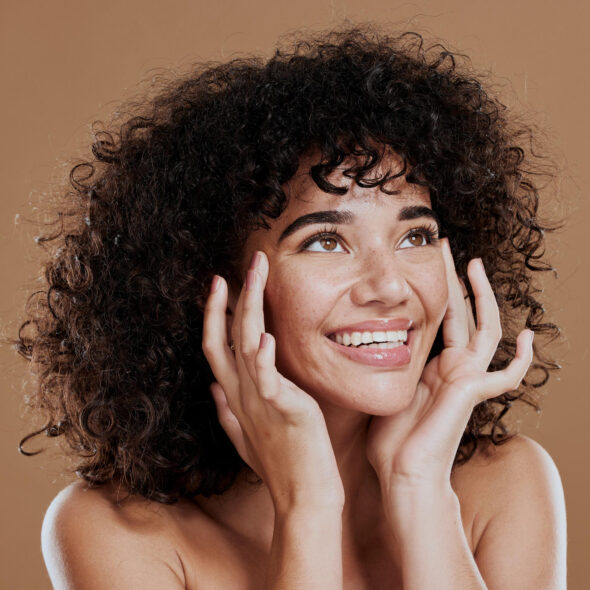 Woman with curly hair smiling brightly while touching her face, expressing joy and confidence.