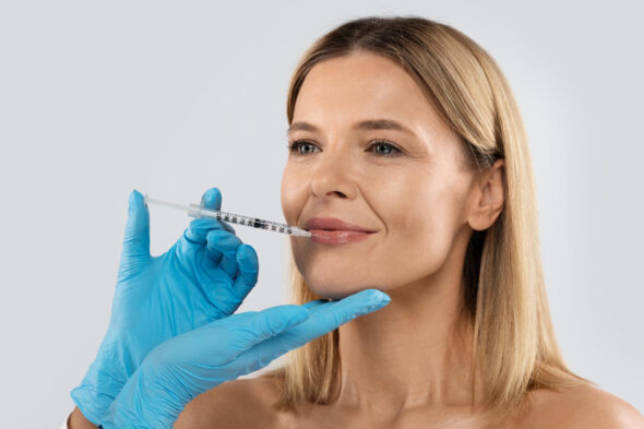 Blue-gloved clinician supports a blonde woman’s chin while holding a syringe near her lips during a cosmetic injection.