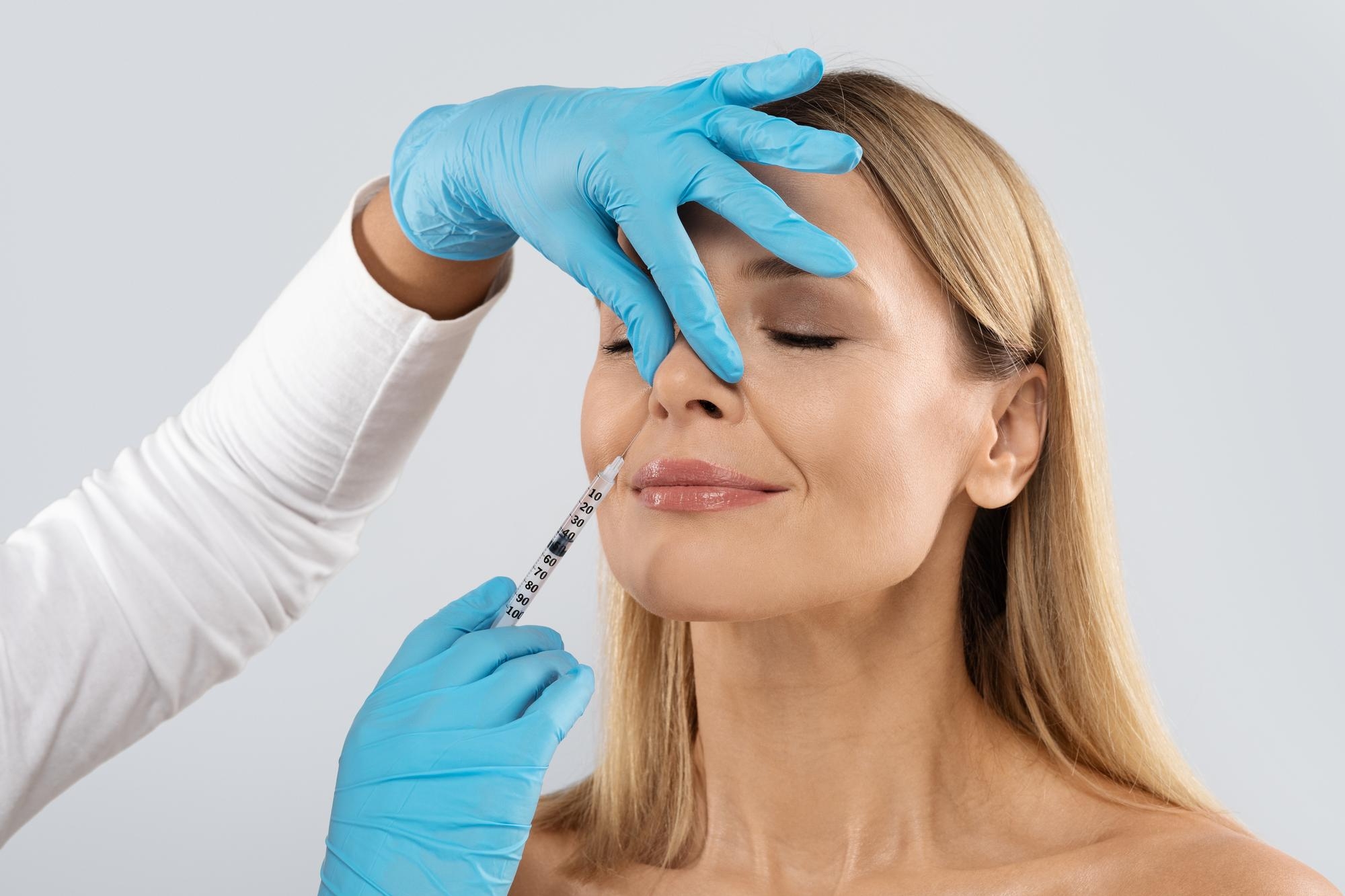 Woman with eyes closed receiving a lip filler injection from a practitioner wearing blue medical gloves.