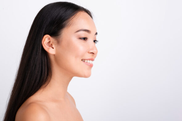Side-profile of a smiling woman with long dark hair and radiant skin against a light background.