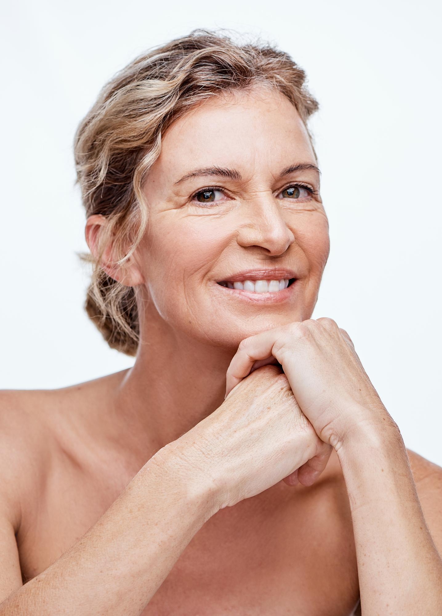 Smiling mature woman rests her chin on clasped hands against a white background.