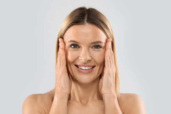 Smiling woman gently pressing her cheeks with both hands, suggesting skin care or facial lifting.