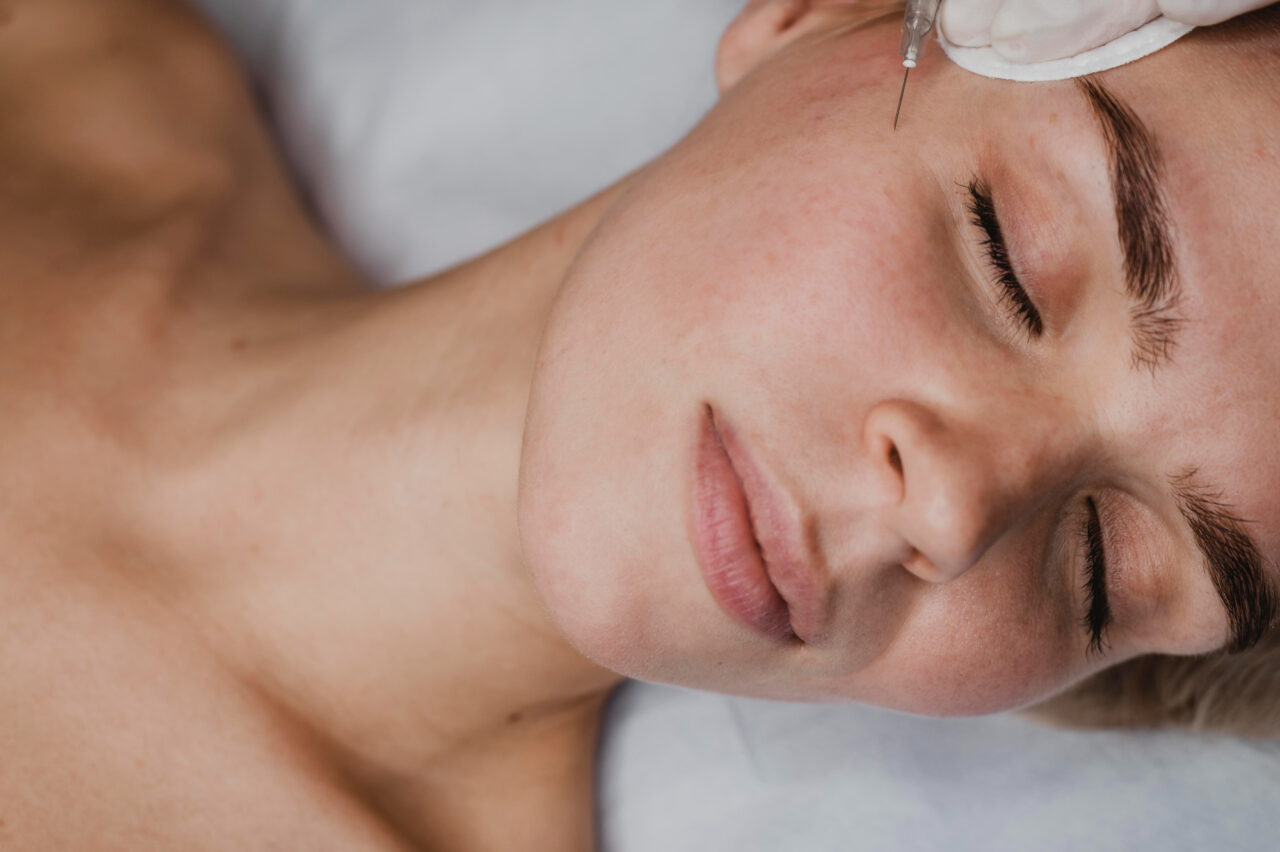 Close-up of a woman lying down with eyes closed while a syringe is positioned under her eye.