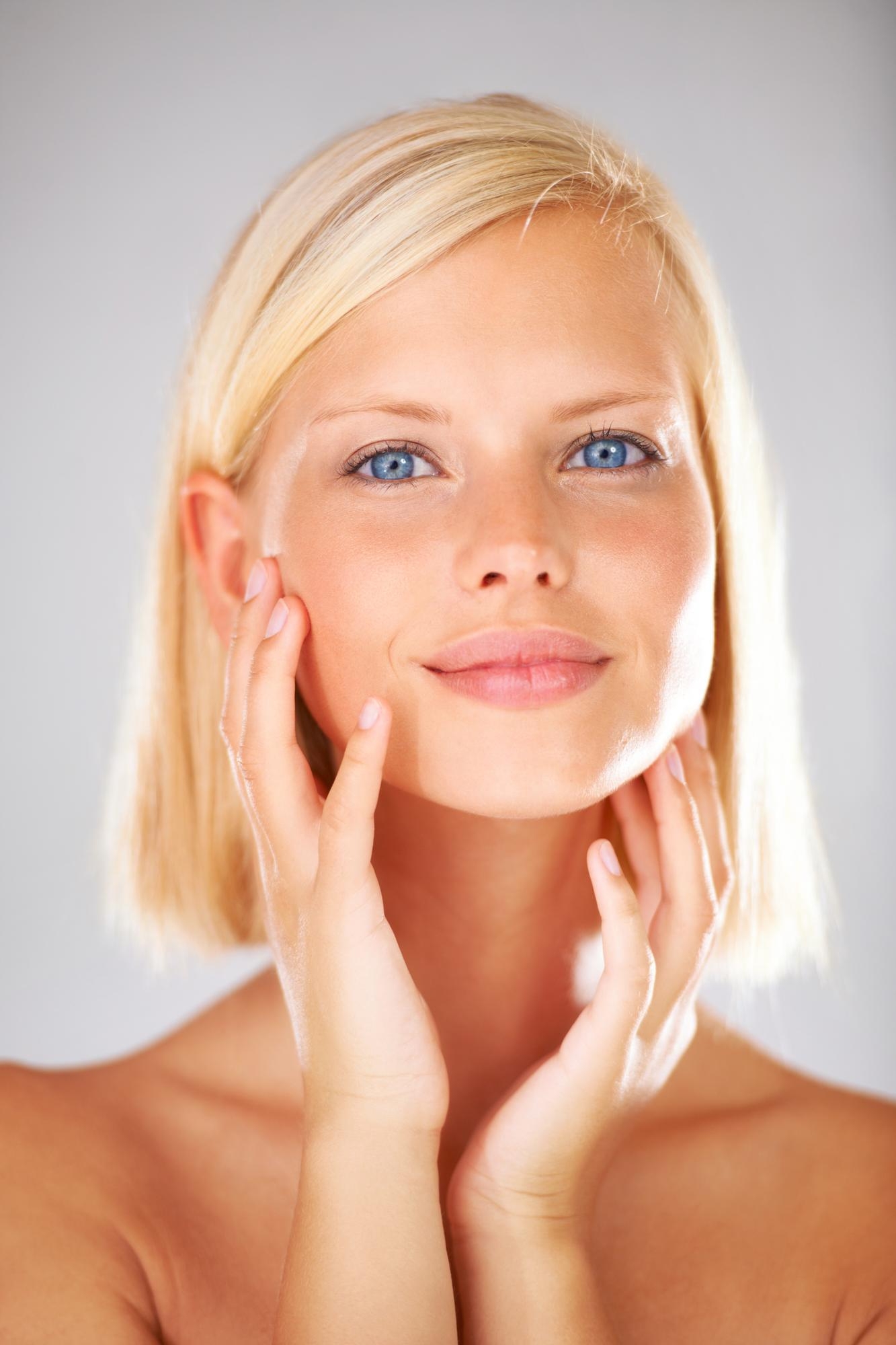 Close-up portrait of a blonde woman with blue eyes gently touching her face against a light gray background.
