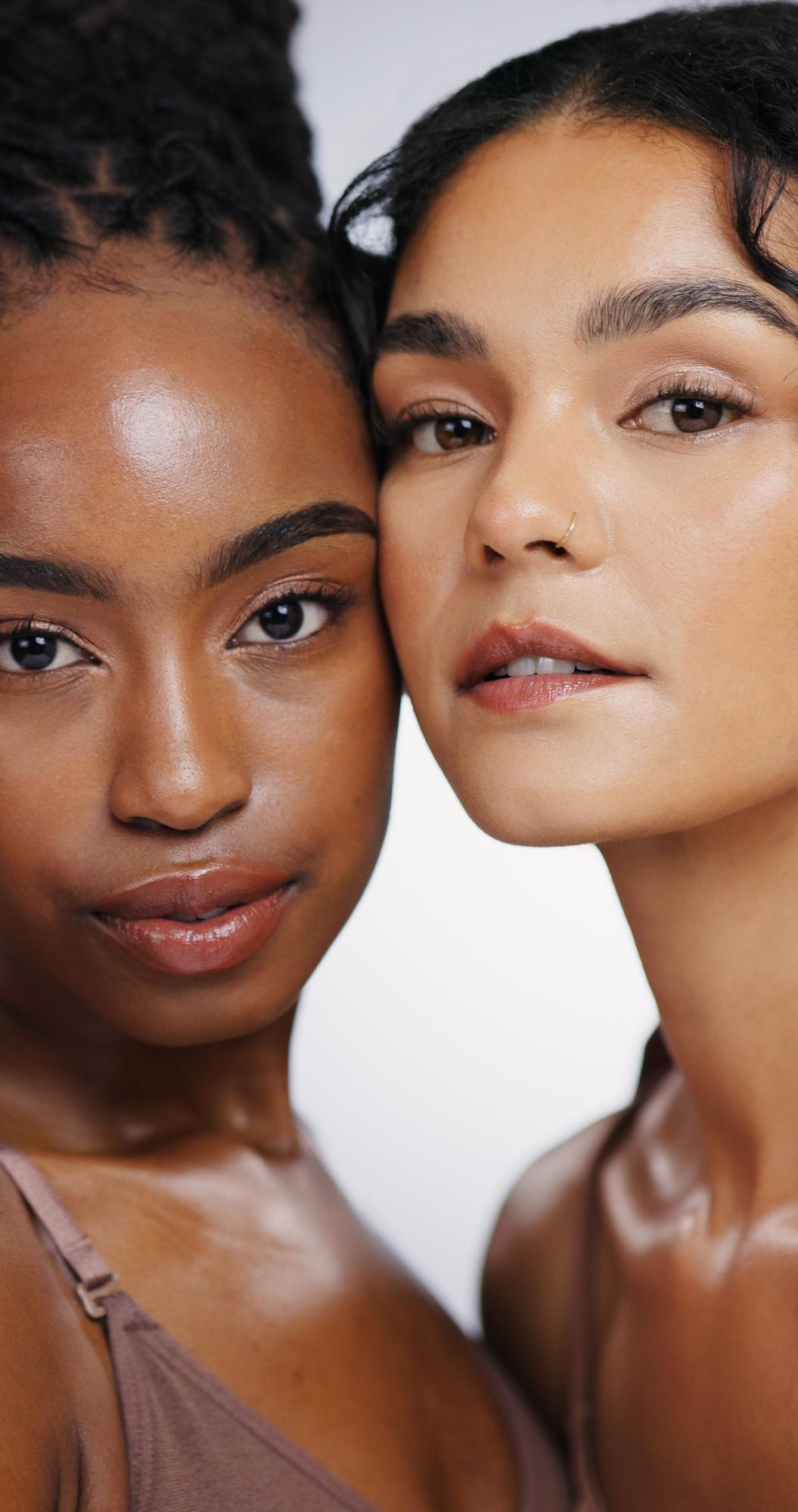 Two people standing close together, both with natural makeup and glowing skin, looking directly at the camera against a light background.