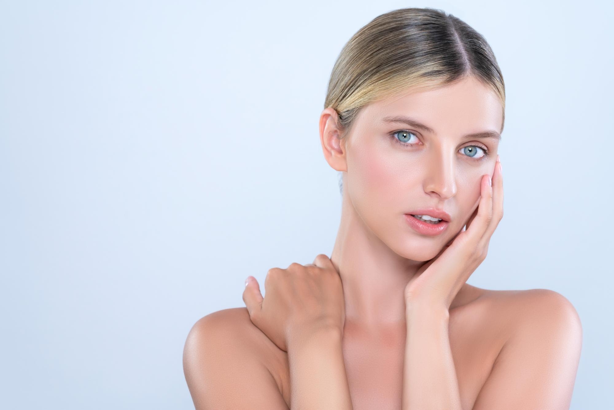 Close-up portrait of a woman with clear glowing skin and blue eyes resting her hand on her cheek against a soft blue background.