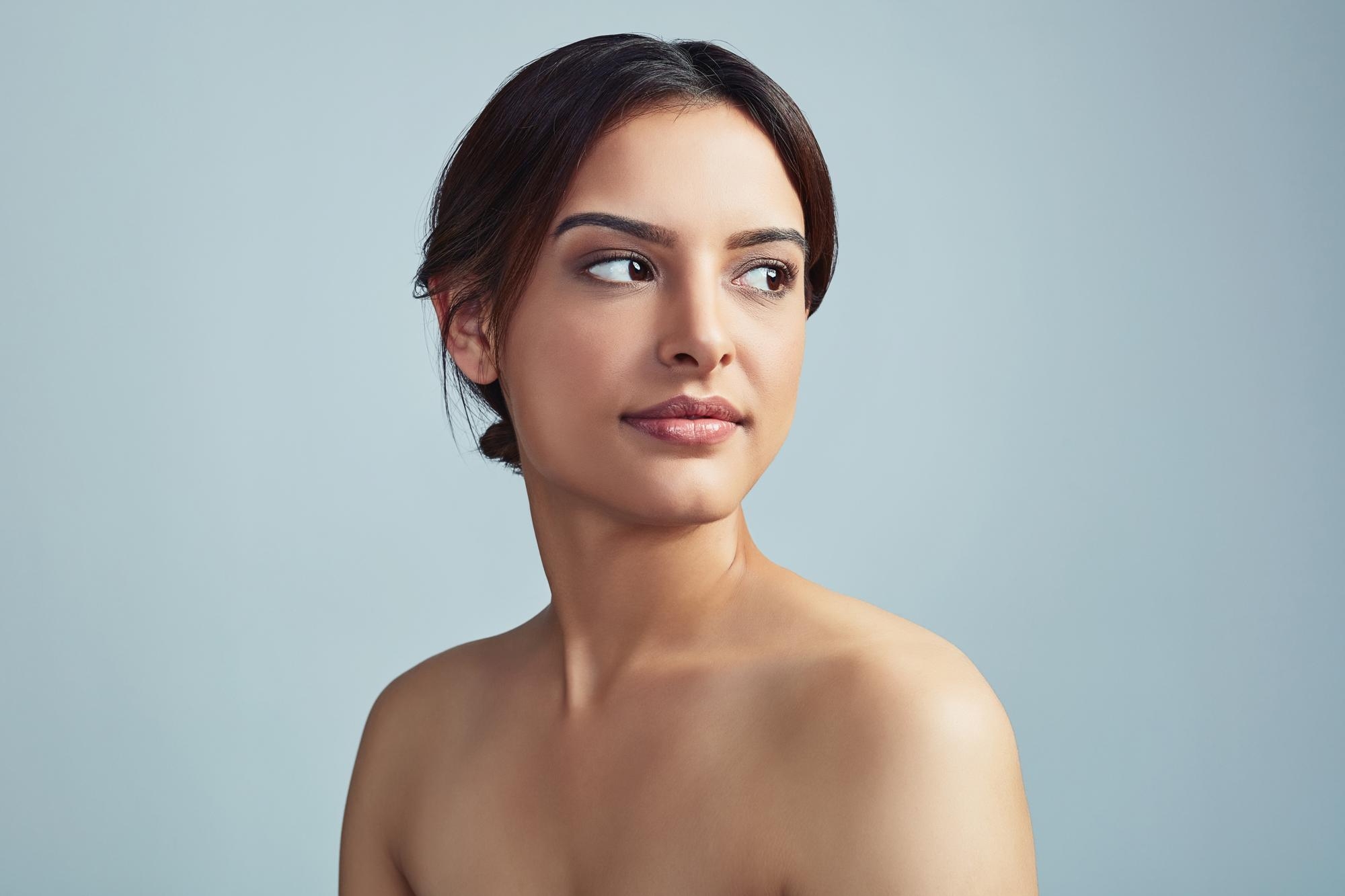 Beauty portrait of a woman with minimal makeup and smooth skin, looking to the side with shoulders bare against a muted backdrop.