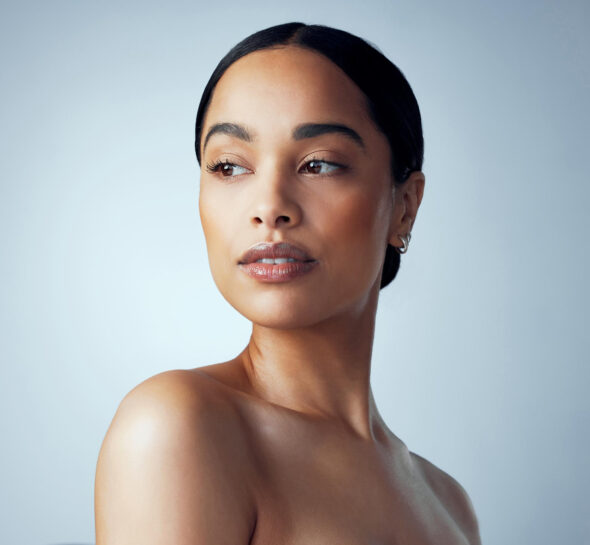 Beauty portrait of a woman with minimal makeup and smooth skin, looking to the side with shoulders bare against a muted backdrop.