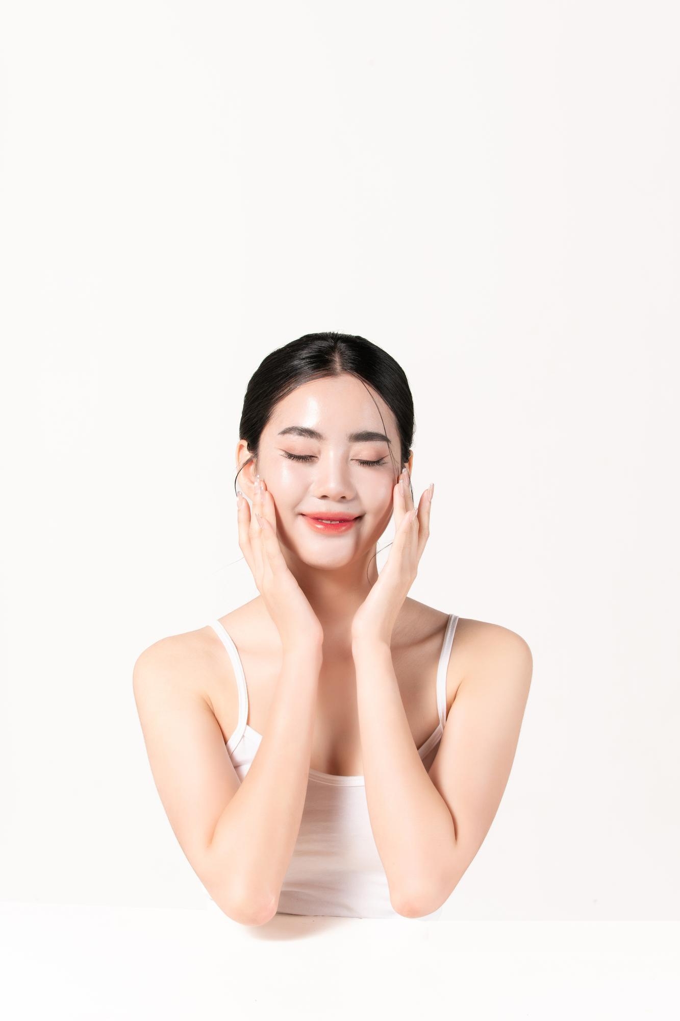 Young Asian woman with eyes closed softly pressing her cheeks with both hands, wearing a white tank top against a clean white background.