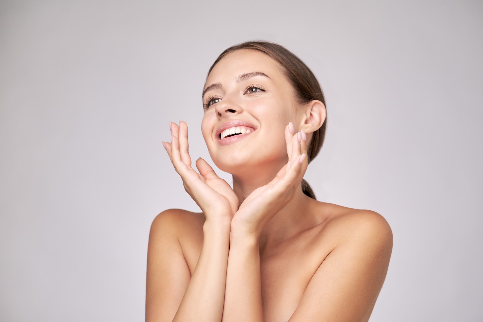 Brunette woman smiling brightly while lifting her face slightly upward and framing her cheeks with both hands against a neutral gray backdrop.