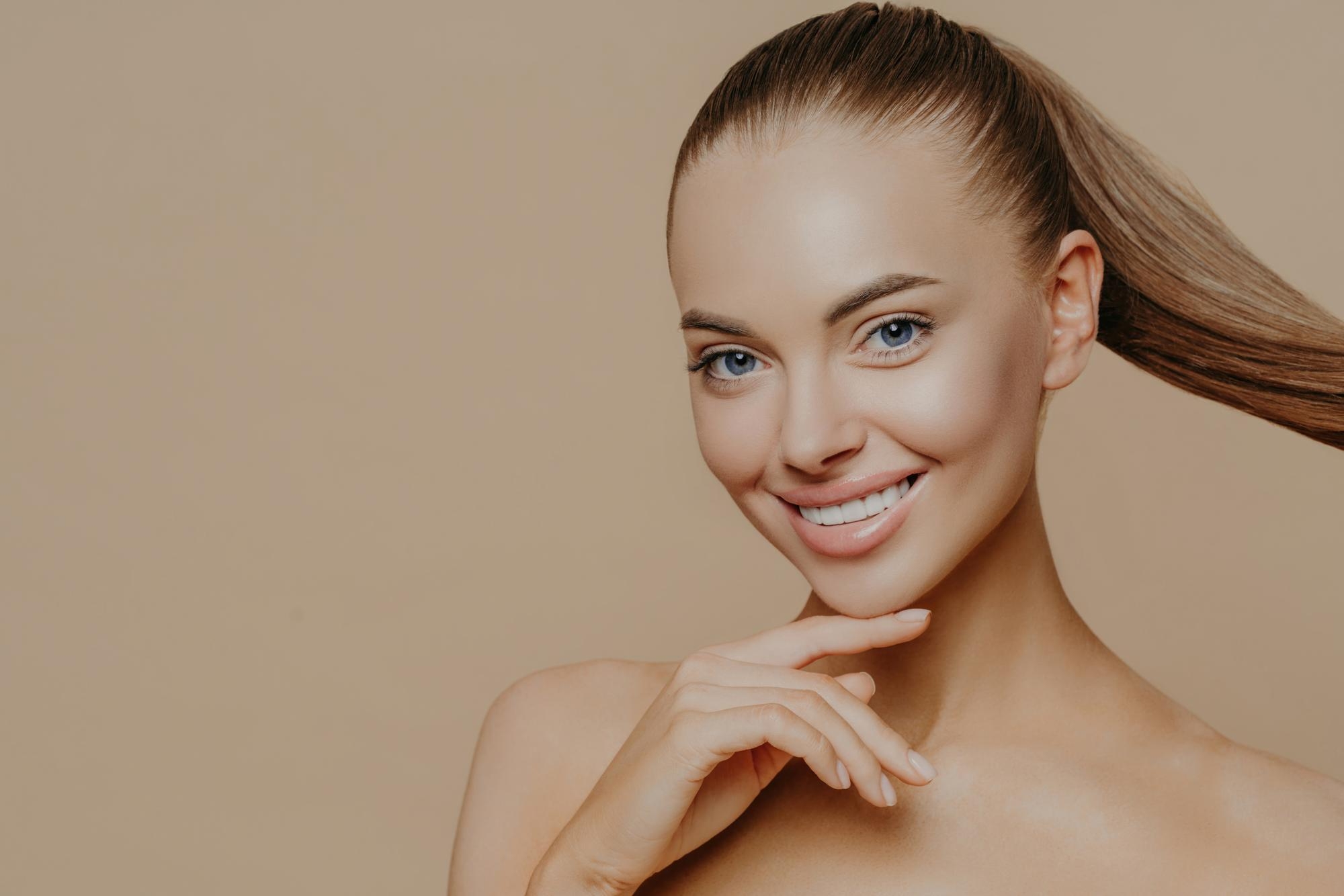 Smiling woman with a sleek ponytail and natural makeup posing against a beige background, highlighting healthy skin.