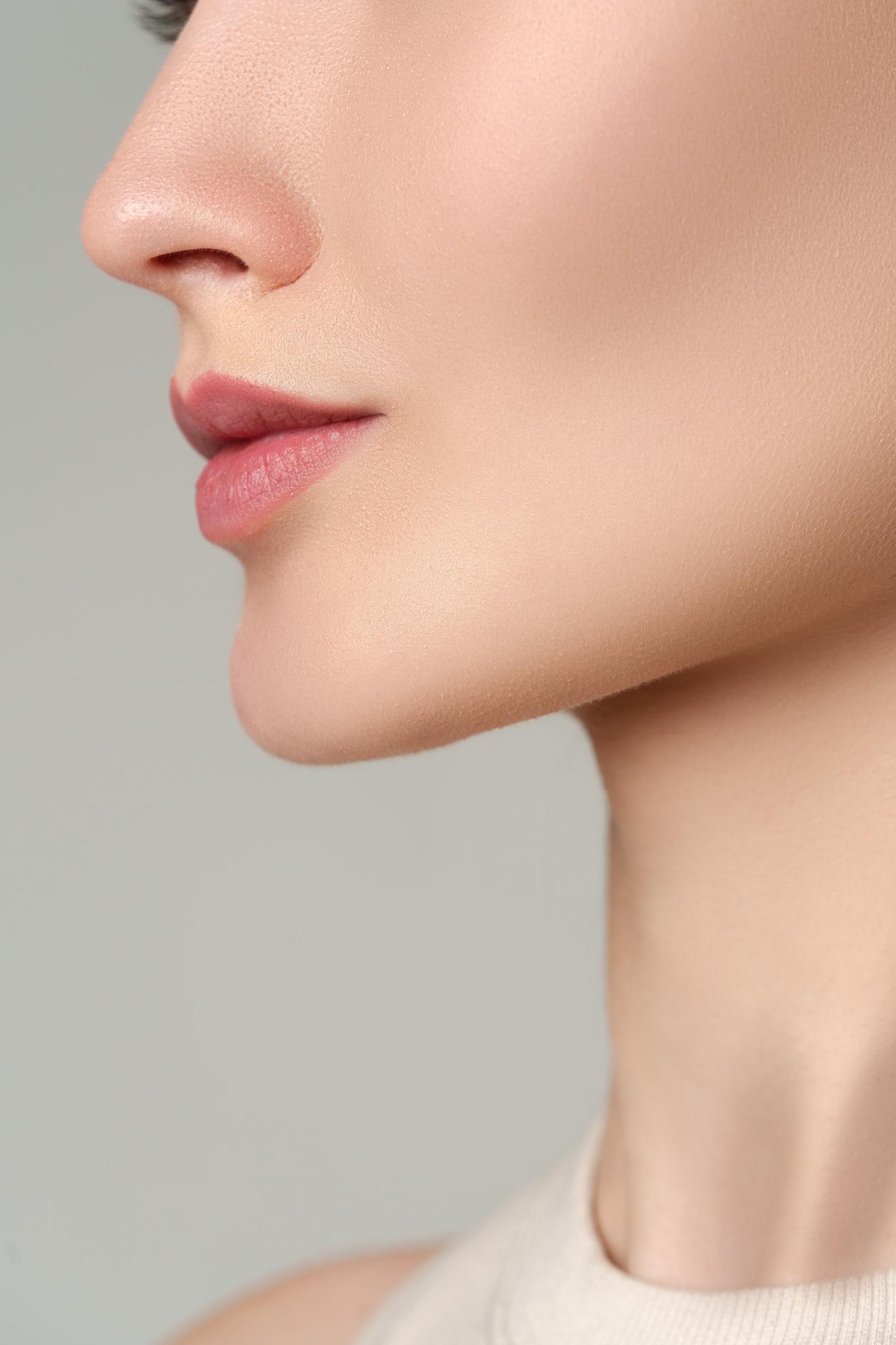Side profile close-up of a woman’s lower face showing smooth skin, natural pink lips, and a defined jawline against a neutral background.