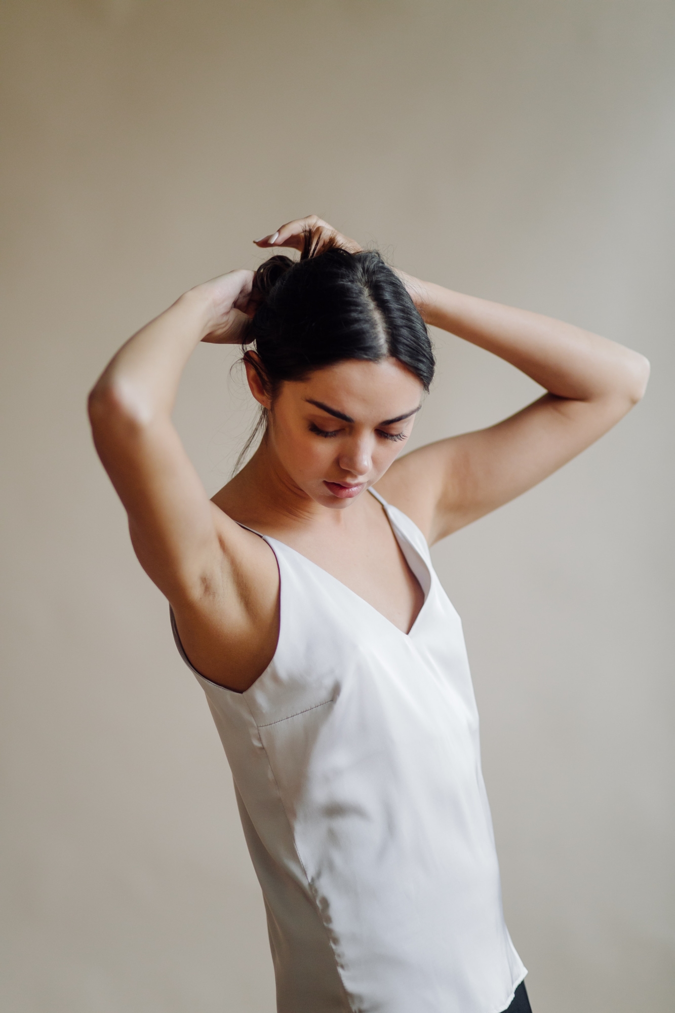Woman in a sleeveless top tying her hair up with arms raised, revealing smooth underarms.