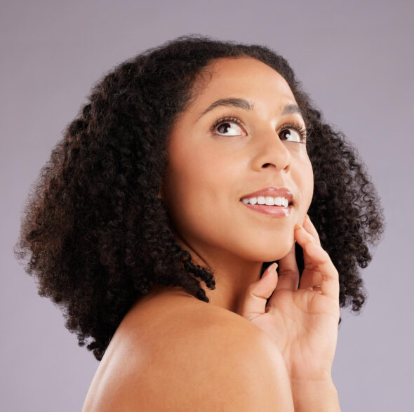 Woman with natural curly hair looking upward while softly touching her chin, set against a neutral backdrop.