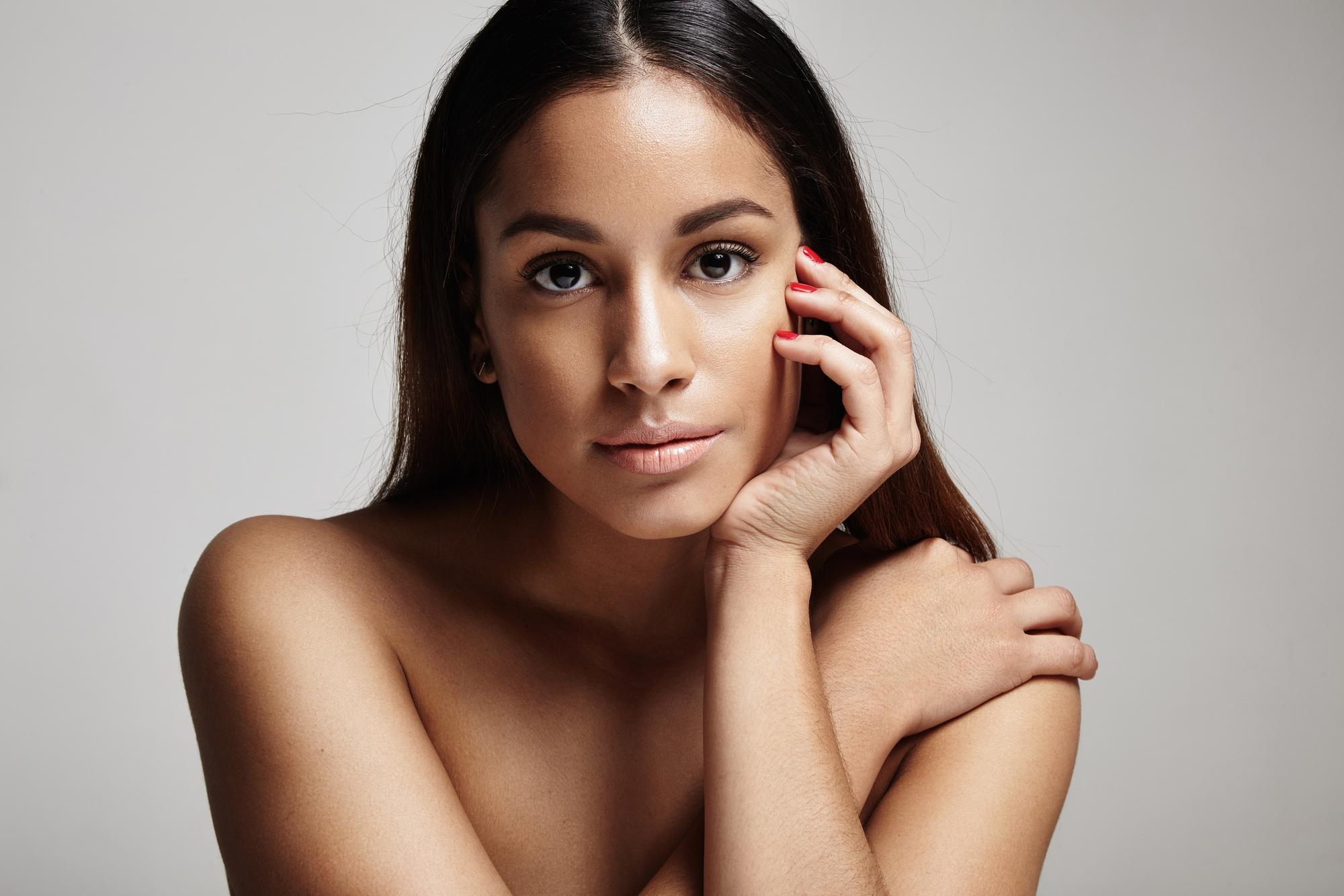 Woman with wavy brown hair tilting her head slightly while touching her neck, wearing subtle jewelry.
