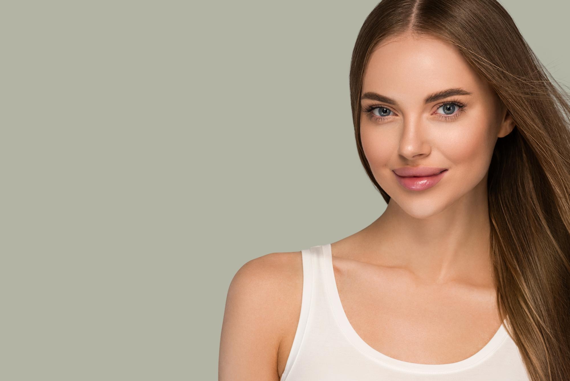 Studio portrait of a young woman with clear skin and long brown hair, smiling softly against a neutral background.